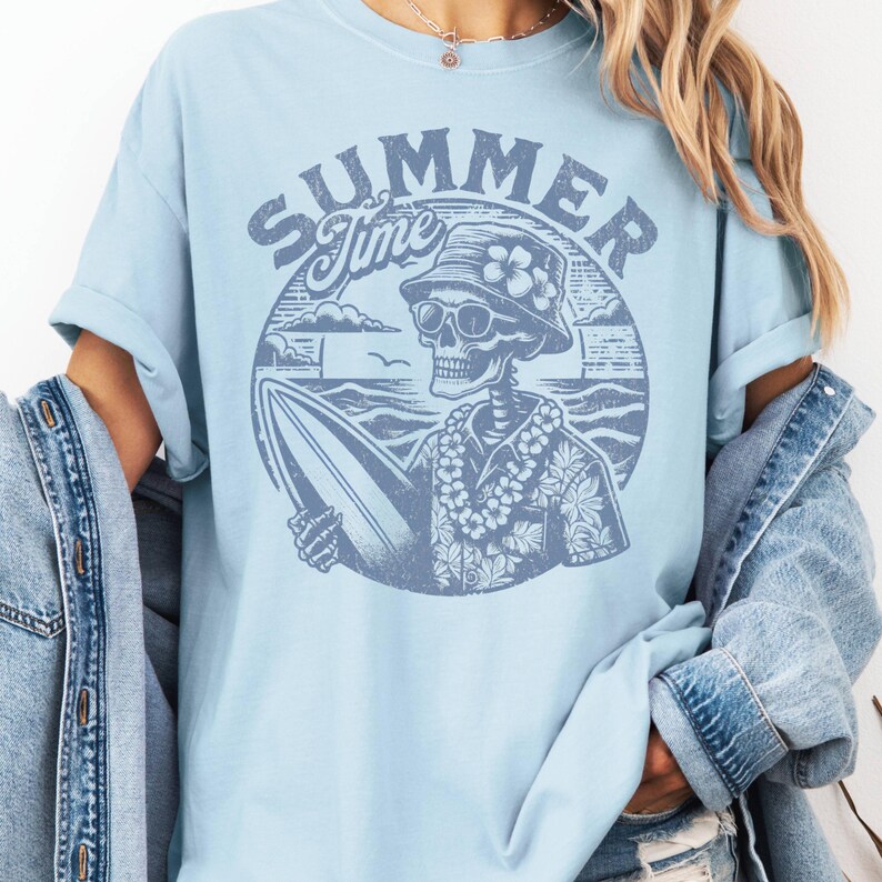 May include: Light blue t-shirt with a graphic of a skeleton surfer wearing sunglasses, a floral lei, and a bucket hat. The design includes the words "SUMMER Time" in a circular design with ocean waves and clouds.