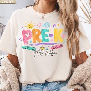 May include: An ivory t-shirt featuring a colorful graphic with the text PRE-K and Mrs. Wilson in black script. The design includes crayon-style lettering in pink, orange, teal, and yellow, surrounded by a sun, hearts, stars, and two crayons.