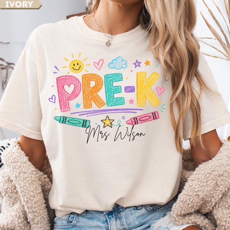 May include: An ivory t-shirt featuring a colorful graphic with the text PRE-K and Mrs. Wilson in black script. The design includes crayon-style lettering in pink, orange, teal, and yellow, surrounded by a sun, hearts, stars, and two crayons.