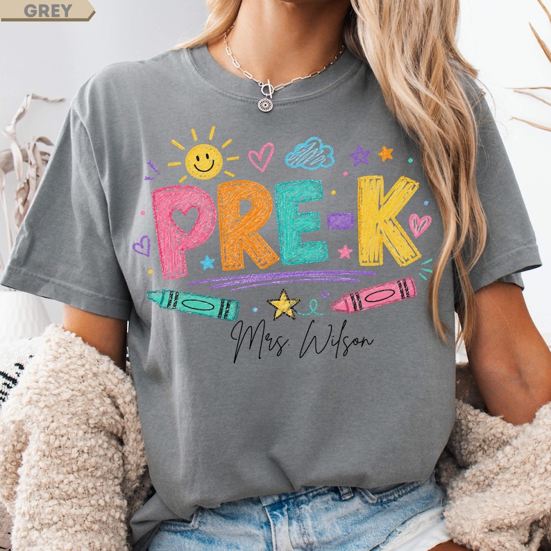 May include: A grey t-shirt featuring the text PRE-K in colorful, crayon-textured letters. The design includes a yellow sun, hearts, stars, a cloud, and two crayons. Below the text, the name Mrs. Wilson is printed in a black cursive script font.