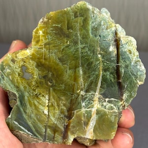 May include: A raw, polished mineral specimen featuring a vibrant olive-green and yellow colour palette. The stone displays intricate, swirling patterns, translucent sections, and distinct dark brown mineral veins running vertically across the surface.