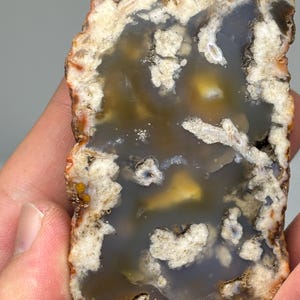 May include: A rectangular agate stone with a polished surface and natural patterns. The stone displays a mix of colours, including grey, brown, yellow, and white. The edges are rough and textured, while the interior has a smooth, swirling design.