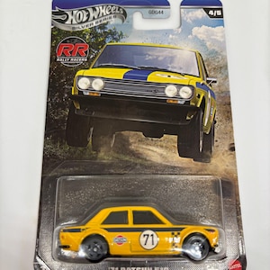 May include: A yellow and blue '71 Datsun 510 die-cast toy car from the Hot Wheels Silver Series. The car features black wheels, a racing stripe, and the number 71 on the side. The packaging includes a picture of the car in action.