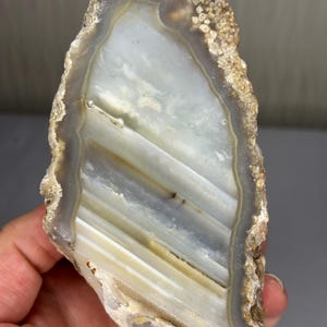 Op de afbeelding: Een gepolijste agaatplak met doorschijnende witte en grijze horizontale banden. De steen heeft een ruwe, getextureerde en onregelmatige buitenrand met aardse bruine en beige tinten, die contrasteert met het gladde, glanzende binnenoppervlak.
