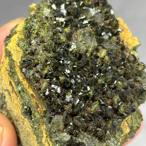 Può includere: Un esemplare minerale grezzo caratterizzato da un ammasso di cristalli scuri, metallici e sfaccettati incastonati in una matrice di roccia verde oliva brillante e giallo senape. La superficie mostra una densa concentrazione di punte cristalline.