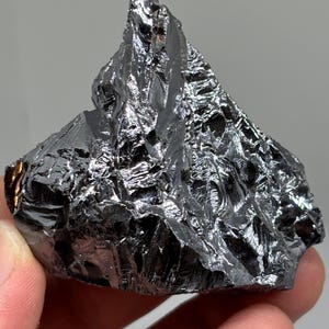 Könnte beinhalten: Ein dunkles, reflektierendes Mineralpräparat mit unregelmäßiger Form. Die Oberfläche des Gesteins ist stark strukturiert mit scharfen Kanten und metallischem Glanz. Die Farbe ist ein tiefes, glänzendes Schwarz mit silbernen Reflexen.