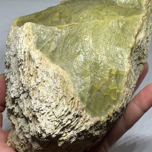 May include: A large, raw mineral specimen featuring a smooth, translucent, pale yellow-green crystalline surface contrasted by a rough, textured, beige-colored outer crust with layered, flaky formations across the exterior of the stone.