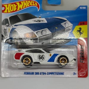 May include: A white and blue Ferrari 365 GTB4 Competizione die-cast toy car with gold wheels. The car has the number "66" on the side and red and blue accents. The packaging includes the Hot Wheels logo and the Ferrari logo.