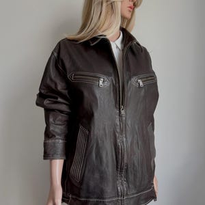 May include: A dark brown leather jacket with a zip-up front, two chest pockets, and two side pockets. The jacket features white stitching and is displayed on a mannequin. The jacket is a medium length and has a classic design.