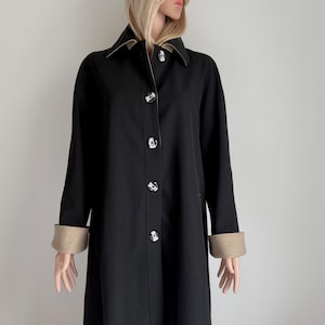 Può includere: Un cappotto lungo nero con colletto e polsini beige a contrasto. Il cappotto presenta bottoni decorativi ed è abbinato a un cappello nero. Uno stile classico, adatto a varie occasioni.