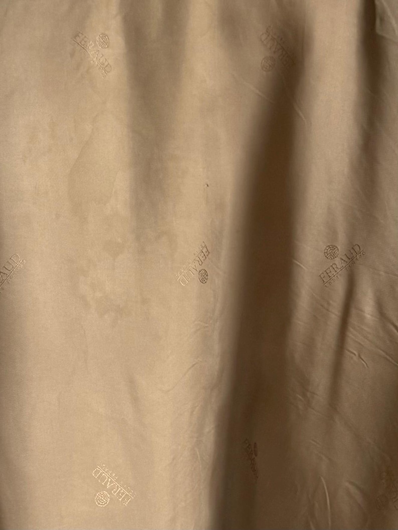 May include: A light tan fabric featuring a repeating pattern of the word FERAUD printed in a darker tan shade. The text is accompanied by a small circular logo above the brand name, creating a subtle, monochromatic design across the material.