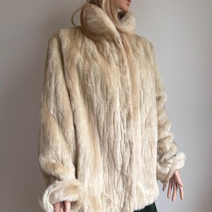 May include: A cream-colored faux fur jacket featuring a high collar, long sleeves with cuffed wrists, and a textured, vertical striped pattern throughout the garment. The jacket is worn over a dark green skirt with a subtle shimmering finish.
