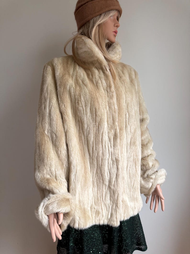 May include: A cream-colored faux fur jacket featuring a high collar, long sleeves with cuffed wrists, and a textured, vertical striped pattern throughout the garment. The jacket is worn over a dark green skirt with a subtle shimmering finish.
