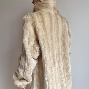May include: A cream-colored faux fur jacket with a high collar and long sleeves, featuring a vertical striped texture throughout the garment. The jacket is worn with a brown beanie hat and a dark green skirt visible at the bottom hem.