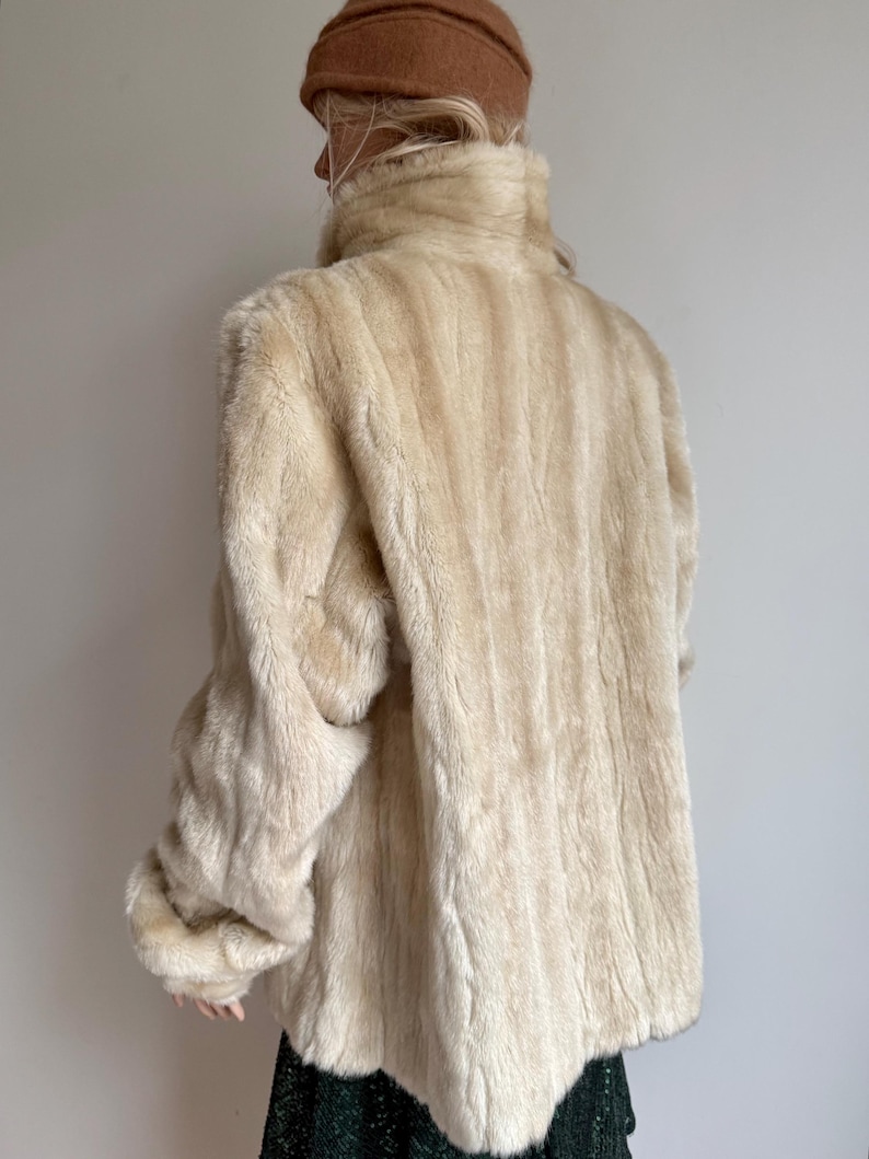 May include: A cream-colored faux fur jacket with a high collar and long sleeves, featuring a vertical striped texture throughout the garment. The jacket is worn with a brown beanie hat and a dark green skirt visible at the bottom hem.