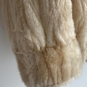 May include: A cream-colored faux fur coat featuring a long-pile texture and a thick, banded cuff at the sleeve. The garment displays a soft, dense appearance with a consistent light beige hue throughout the visible fabric surface.