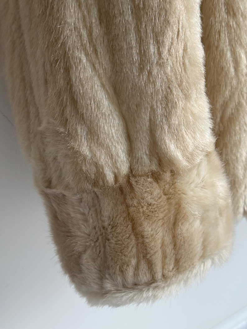 May include: A cream-colored faux fur coat featuring a long-pile texture and a thick, banded cuff at the sleeve. The garment displays a soft, dense appearance with a consistent light beige hue throughout the visible fabric surface.