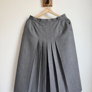 May include: A gray pleated skirt hanging on a wooden hanger. The skirt has a high waist and a knee-length hem. The fabric appears to be a medium-weight wool or wool blend. The skirt is hanging against a white wall.