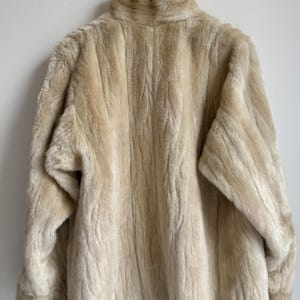 May include: A cream-colored faux fur jacket with a high collar and long sleeves. The garment features a textured, vertical striped pattern throughout the plush material, providing a consistent, soft appearance across the entire outer surface.