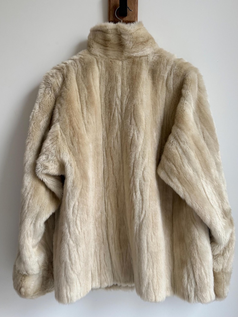 May include: A cream-colored faux fur jacket with a high collar and long sleeves. The garment features a textured, vertical striped pattern throughout the plush material, providing a consistent, soft appearance across the entire outer surface.