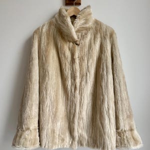 May include: A cream-colored faux fur coat featuring a high collar, long sleeves with flared cuffs, and a single button closure at the neckline. The textured material has a vertical, wavy pattern throughout the entire garment.