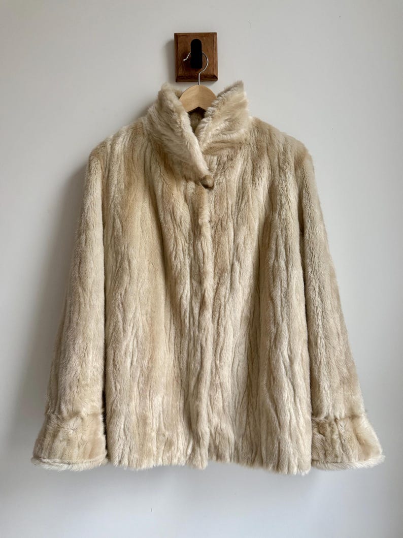 May include: A cream-colored faux fur coat featuring a high collar, long sleeves with flared cuffs, and a single button closure at the neckline. The textured material has a vertical, wavy pattern throughout the entire garment.