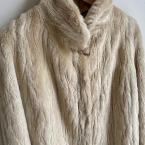 May include: A cream-colored faux fur coat with a high collar and a single large button closure at the neck. The coat features a textured, wavy pile throughout the fabric and is displayed on a wooden hanger against a plain white background.