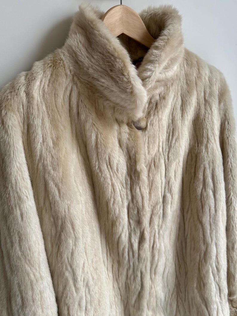 May include: A cream-colored faux fur coat with a high collar and a single large button closure at the neck. The coat features a textured, wavy pile throughout the fabric and is displayed on a wooden hanger against a plain white background.