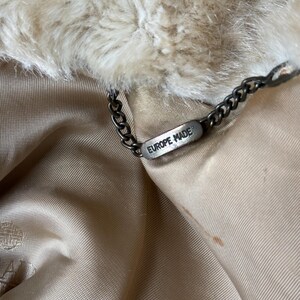 May include: A beige faux fur collar attached to a smooth, light tan satin lining. A dark metal chain is connected to a small, rectangular metal tag engraved with the text EUROPE MADE, which hangs from the inner neckline of the garment.