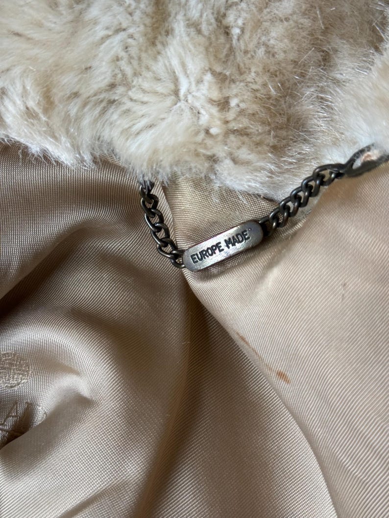 May include: A beige faux fur collar attached to a smooth, light tan satin lining. A dark metal chain is connected to a small, rectangular metal tag engraved with the text EUROPE MADE, which hangs from the inner neckline of the garment.