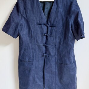 Könnte beinhalten: Marineblaue Jacke mit kurzen Ärmeln, V-Ausschnitt und drei Knebelverschlüssen vorne. Die Jacke hat zwei Vordertaschen und ein kleines Etikett mit der Aufschrift "cob" auf der Innenseite. Die Jacke hängt an einem weißen Kleiderbügel.