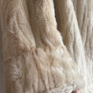 May include: A long-sleeved coat made of cream-colored faux fur. The garment features a thick, textured pile with a soft appearance and a wide cuff detail at the end of the sleeve, showcasing the dense, plush quality of the synthetic material.