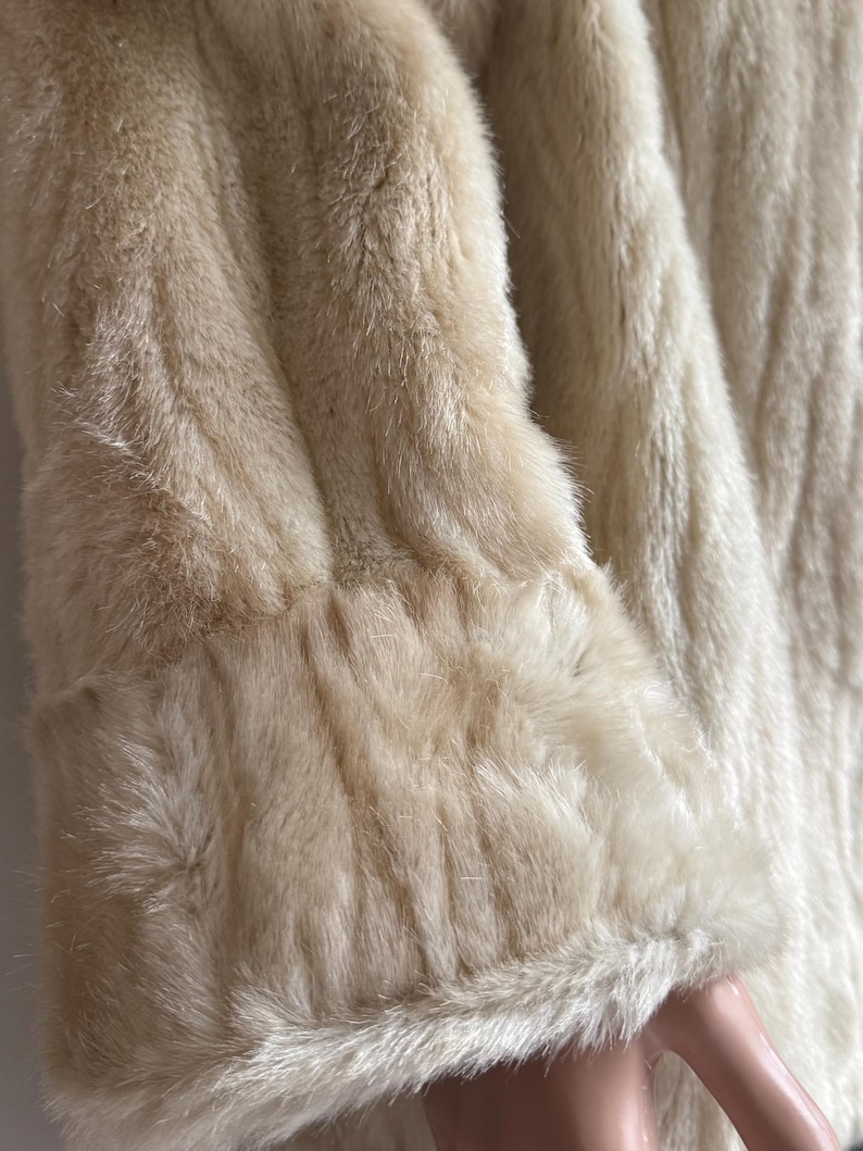 May include: A long-sleeved coat made of cream-colored faux fur. The garment features a thick, textured pile with a soft appearance and a wide cuff detail at the end of the sleeve, showcasing the dense, plush quality of the synthetic material.