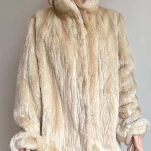 May include: A cream-colored faux fur coat with a high collar and long sleeves. The coat features a textured, vertical striped pattern throughout the fabric, with folded cuffs at the ends of the sleeves and a straight hemline that falls at the hip.