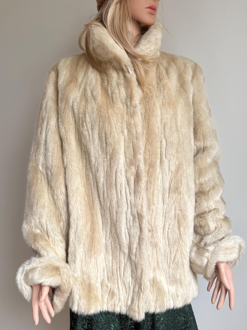 May include: A cream-colored faux fur coat with a high collar and long sleeves. The coat features a textured, vertical striped pattern throughout the fabric, with folded cuffs at the ends of the sleeves and a straight hemline that falls at the hip.