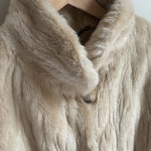 May include: A cream-colored faux fur coat featuring a wide collar and a single large, round, marbled button closure at the neckline. The coat has a soft, textured pile and is displayed on a light-colored wooden hanger against a plain background.
