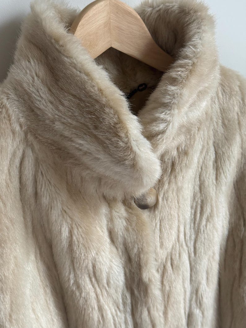 May include: A cream-colored faux fur coat featuring a wide collar and a single large, round, marbled button closure at the neckline. The coat has a soft, textured pile and is displayed on a light-colored wooden hanger against a plain background.