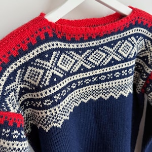 May include: A navy blue sweater with a red neckline and cuffs. The sweater features a white geometric pattern across the chest and sleeves. The sweater is hanging on a white hanger.