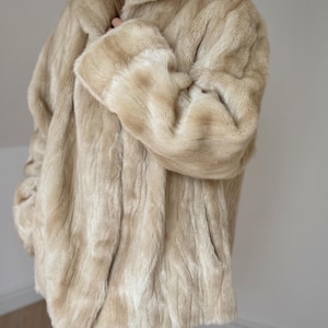 May include: A beige faux fur coat with a high collar and long sleeves. The coat features a soft, textured pile with vertical variations in color, ranging from light cream to tan, and is worn over blue denim jeans, showing a hip-length cut.