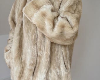 May include: A beige faux fur coat with a high collar and long sleeves. The coat features a soft, textured pile with vertical variations in color, ranging from light cream to tan, and is worn over blue denim jeans, showing a hip-length cut.