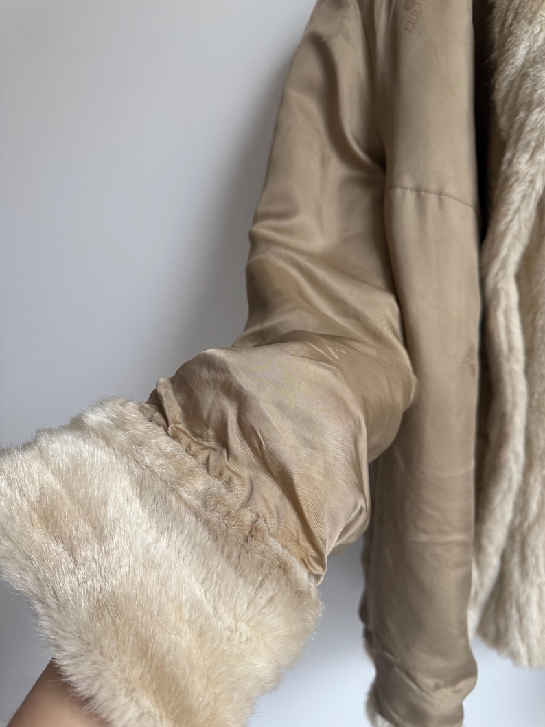 May include: A beige jacket featuring a smooth satin-like finish on the exterior and a plush, cream-colored faux fur lining. The sleeve ends in a wide, thick cuff made of the same soft, cream-colored faux fur material, providing a textured contrast.