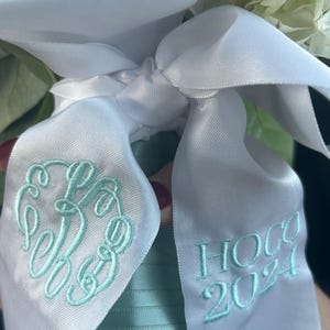May include: White satin ribbon tied in a bow, embroidered with a monogram and "HOCO 2024" in light blue. The ribbon is part of a bouquet with white flowers and green foliage.