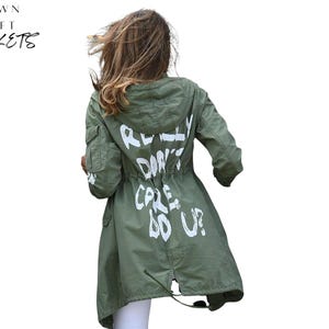 Könnte beinhalten: Olivgrüne Kapuzenjacke mit weißem Text "REALLY DON'T CARE DO U?". Die Jacke hat einen Kordelzug in der Taille und Taschen. Der Markenname "CROWN CRAFT JACKETS" befindet sich oben links.