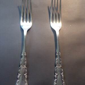 May include: Two silver forks with ornate, detailed handles. Each fork has four tines and a decorative pattern on the handle. The forks are displayed against a dark background, highlighting their polished finish.
