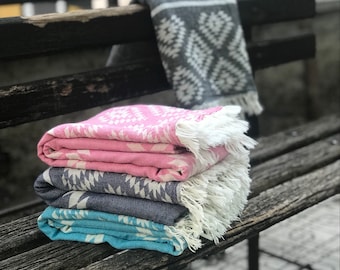 May include: A stack of four folded, patterned blankets in shades of pink, navy, and teal, with white fringe. A gray blanket with a white geometric design is draped over a wooden bench. The blankets have a geometric pattern.