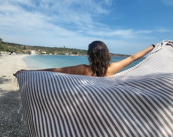 Puede incluir: Una persona de pie en una playa sosteniendo una toalla rayada blanca y marrón frente a un cielo azul y el océano.
