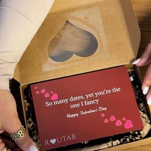 Könnte beinhalten: Eine Valentinstags-Geschenkbox mit herzförmigem Fenster. Im Inneren ist eine rote Karte mit der Aufschrift "So many dates, yet you're the one I fancy. Happy Valentine's Day" zu sehen. Die Box ist braun.