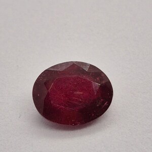 Puede incluir: Una gema de rubí facetada de forma ovalada. La gema de color rojo intenso tiene una superficie pulida con inclusiones internas visibles. El corte de la piedra crea múltiples superficies reflectantes, realzando su brillo. La gema está sobre un fondo claro.