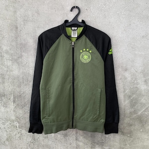Könnte beinhalten: Eine olivgrüne und schwarze Trainingsjacke mit Reißverschluss, die auf der linken Brust das Wappen der deutschen Fußballnationalmannschaft mit vier Sternen darüber zeigt. Die Jacke hat schwarze Ärmel, einen schwarzen Kragen und ein lindgrünes Adidas-Logo.