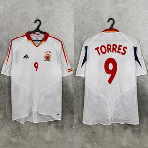 May include: A white Adidas Spain soccer jersey featuring the number 9 in red on the front and back, with the name TORRES printed in red above the number on the back. The shirt includes red and yellow trim on the collar and sleeves.